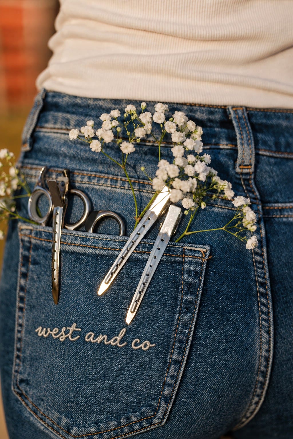 Jeans back pocket with embroidered west and co script, silver shears, a sectioning clip, and baby's breath