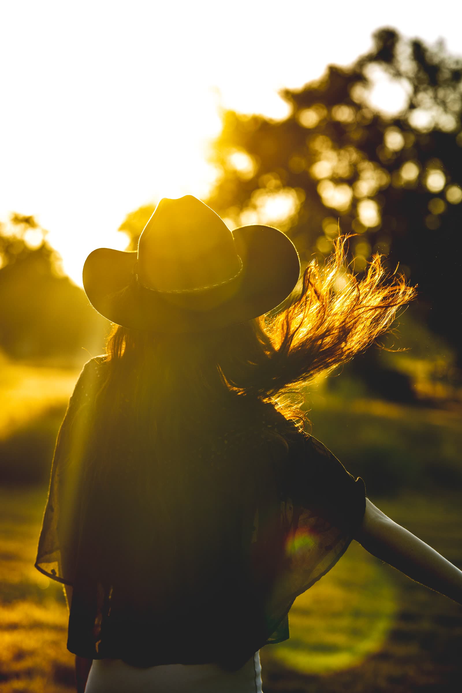 Woman in a cowboy hat, hair catching sunrise light as she turns away in a field
